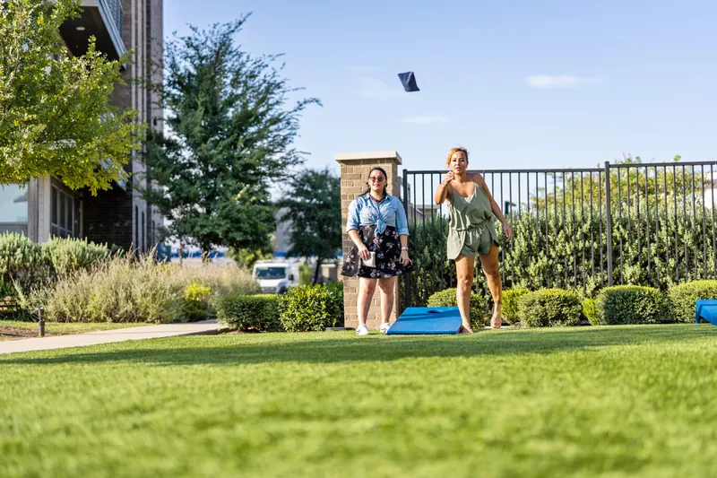 Two women play cornhole on the lawn outside an apartment building