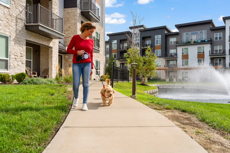 A woman walks a small dog on a path between apartment buildings