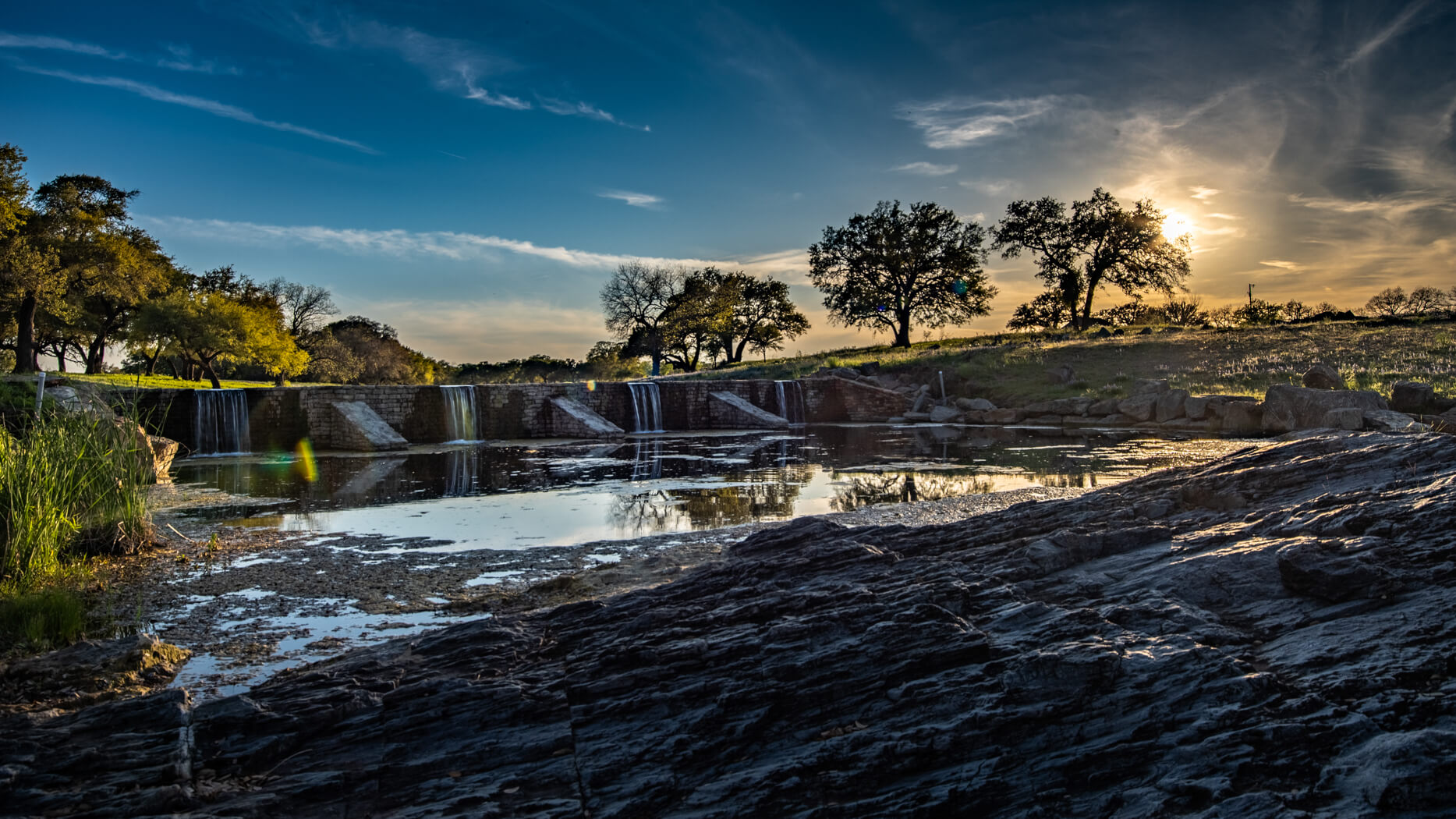 Sunset over a ranch water feature, with trees and open land beyond.